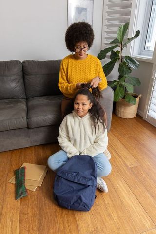 African american mother braiding daughter hair at home preparing for school with backpack