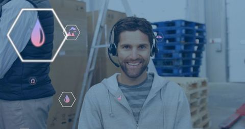 Smiling warehouse operator wearing headset and hoodie near blue pallets with digital hex overlays