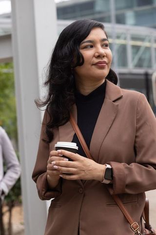 Professional woman holding coffee, wearing blazer and smartwatch, glancing in urban plaza