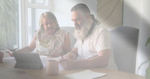 Sunlit mature couple reviewing documents at home desk with tablet and coffee mug