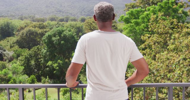 Middle-aged man standing on a balcony, overlooking lush green forested landscape. Ideal for themes of relaxation, tranquility, enjoying nature, outdoor activities, and wellness. Could work well for promoting environmental conservation, vacation spots, mindfulness practices, or outdoor lifestyle.