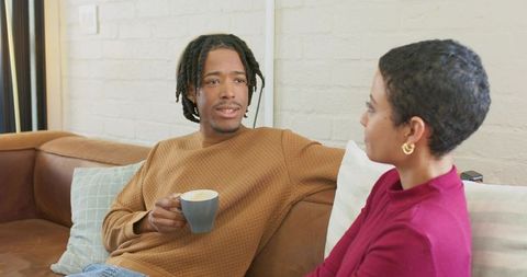 Diverse young couple sipping coffee and chatting on leather sofa, cozy living room moment