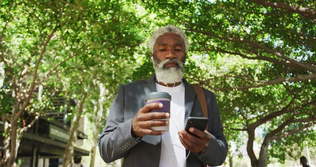 African american senior man holding smartphone drinking coffee while walking in corporate park - Download Free Stock Photos Pikwizard.com