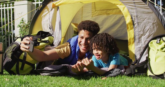 Father and son bonding while having fun in a backyard camping experience. They are lying in a tent and capturing joyful moments with a selfie. This image can be used to portray themes of family bonding, outdoor activities, and joyful parental moments. Perfect for advertisements, family blogs, and outdoor activity promotions.