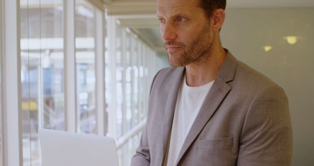 Businessman dressed in blazer using laptop while standing near large office windows. Ideal for workplace, success, technology, modern business, corporate themes.