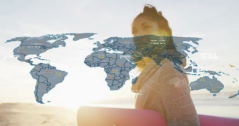 Woman standing holding yoga mat wearing headphones on sunlit beach with world map overlay