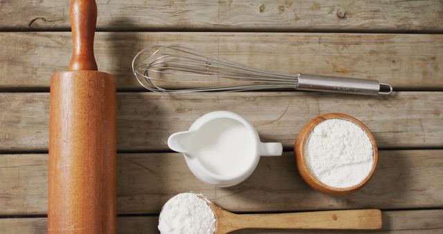 Baking basics are arranged neatly on a rustic wooden table, creating a charming and nostalgic setting. A wooden rolling pin, a metal whisk, a measuring cup with milk, a wooden bowl of flour, and a wooden spoon with flour showcase essential baking supplies. Suitable for use in cooking blogs, bakery promotions, kitchen-related stores, and food magazines.