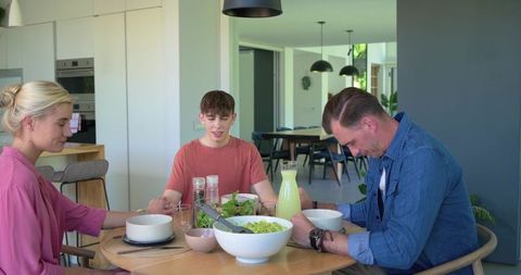 Family holding hands and praying before meal around modern round wooden table