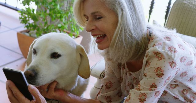 Happy senior caucasian woman with dog using smartphone at home. Retirement, lifestyle, technology, pets and domestic life, unaltered.