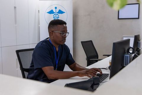 Medical receptionist typing at clinic reception desk wearing navy scrubs and id lanyard