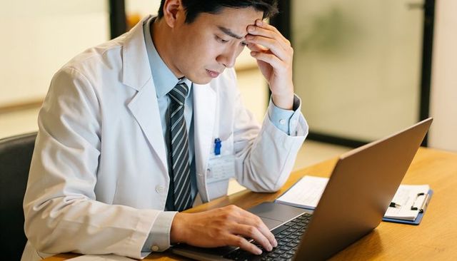 Physician leaning forehead while typing on laptop showing stress and professional burnout