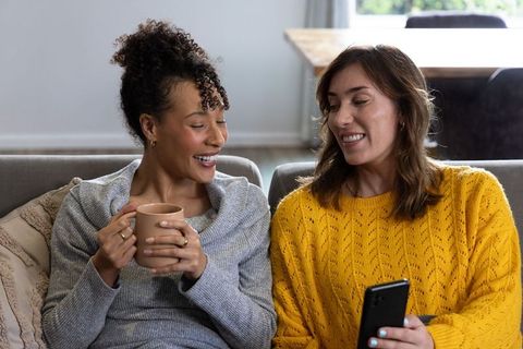 Friends sharing coffee and smartphone on cozy couch, smiling and chatting at home