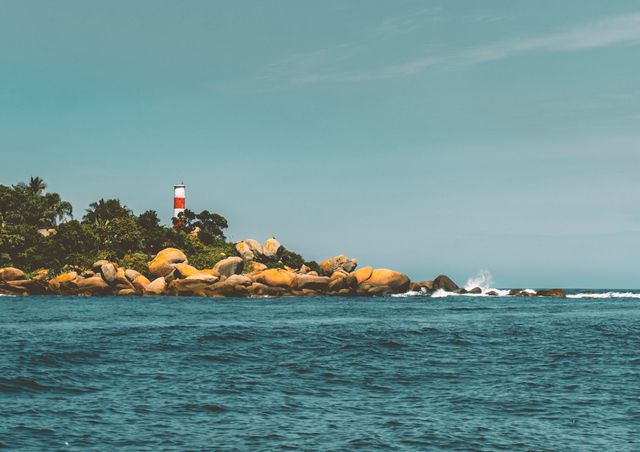 Image showcases a tranquil coastal scene with a lighthouse on a lush, rocky island. Surrounded by the blue ocean, waves gently crash against the rocks under a clear sky. Ideal for themes related to nature, travel, maritime navigation, serenity, and adventure.