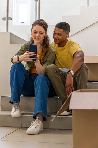 Young couple unpacking in new home sharing smartphone on stairs, casual cozy moving moment