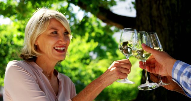 This image shows a smiling mature woman toasting with friends while enjoying an outdoor celebration. The lush green background suggests a warm and pleasant environment. Ideal for use in lifestyle blogs, promotional material for outdoor events, wine and beverage advertisements, or social media campaigns highlighting joyful social gatherings.