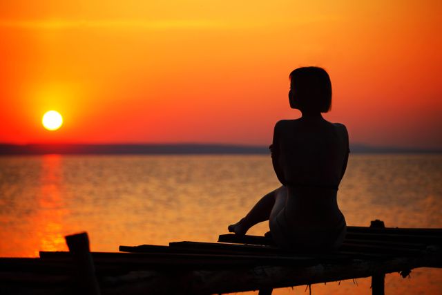 Woman is sitting on a pier during sunset, creating a peaceful and tranquil scene. Ideal for themes of relaxation, serenity, and contemplation. Suitable for use in travel guides, wellness blogs, and inspirational content.