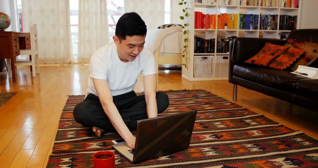 Young man sits on colorful rug in bright, cozy living room, working on laptop. Ideal for remote work concepts, casual lifestyle themes, tech-savvy advertising, home office setups, and interior decoration inspiration.