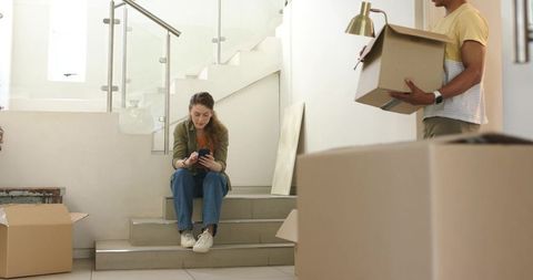 Couple moving into new home on stair landing packing boxes and checking smartphone