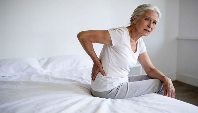 Senior woman sitting on bed holding lower back showing chronic pain and mobility issues