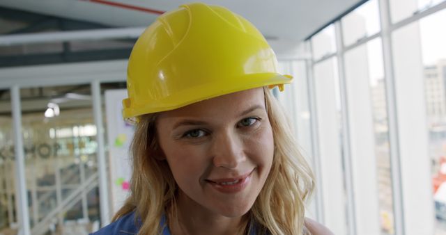 Young female architect wearing a yellow safety helmet, smiling confidently in a modern office with large windows. Ideal for use in promotional materials for construction companies, architecture firms, or safety gear advertisements. Perfect for illustrating topics related to women in construction, workplace diversity, and professional development.