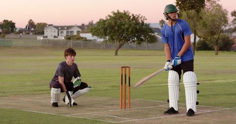 Teenage cricketer batting while wicketkeeper crouching on dusk suburban cricket pitch