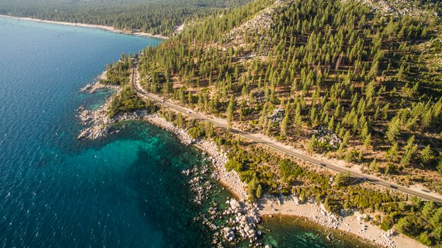 Aerial shot capturing a coastal highway winding through lush forest and rocky coastline with clear turquoise waters. Ideal for travel brochures, environmental campaigns, tourism blogs, and adventure planning materials.