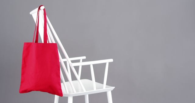 A red tote bag hangs on the back of a white chair against a gray background, with copy space. Its simplicity suggests a minimalist aesthetic or could symbolize eco-friendly shopping habits.