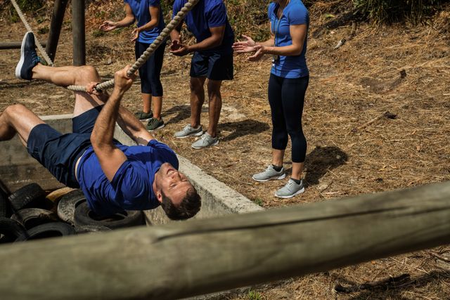 Fit man crossing rope during an obstacle course while team members cheer him on. Ideal for use in fitness, teamwork, and motivational contexts. Perfect for promoting outdoor activities, group training sessions, and athletic events.