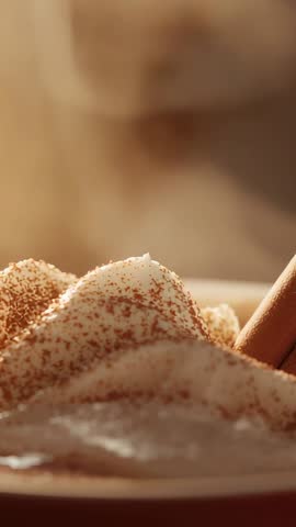 Vertical video steaming cup sending vapor wisps over cocoa-dusted cream with cinnamon stick