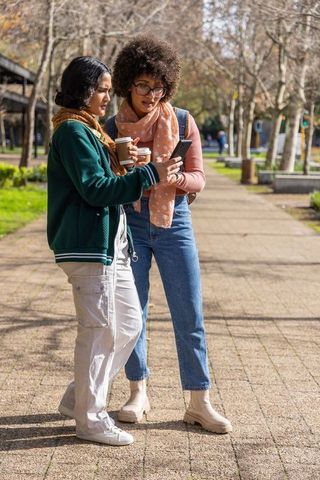 Two female friends holding coffee and showing phone while standing on sunlit park path