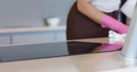 Cleaning glossy induction cooktop with bright pink gloves on modern kitchen countertop