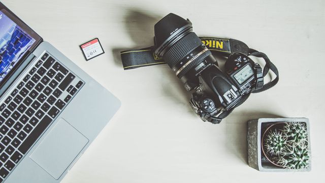 This depicts a clean and organized professional photographer's workspace, featuring a high-end DSLR camera, a laptop, an SD card, and a potted succulent on a wooden desk. Ideal for illustrating concepts related to digital photography, creative workspaces, freelance work, or technology in the creative field.