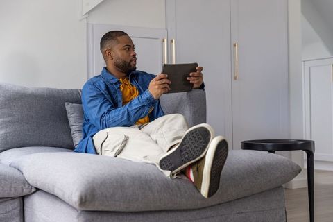 African american man reclining on modern gray sectional sofa using tablet at home for casual leisure