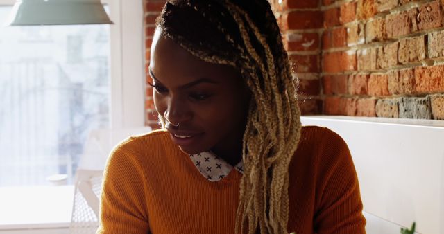 Young woman with dreadlocks relaxing in a cozy, urban cafeteria. She is looking away thoughtfully, dressed in casual attire with an exposed brick wall in the background. This image can be used to represent modern lifestyle, leisure time, or contemporary urban settings. Ideal for articles or advertisements related to coffee shops, lifestyle blogs, or articles focused on relaxation and contemplation.