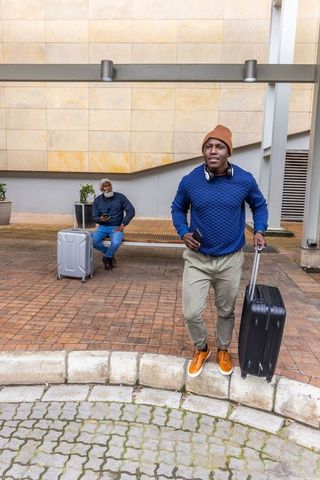 African american traveler pulling suitcase and checking phone at urban curbside pickup