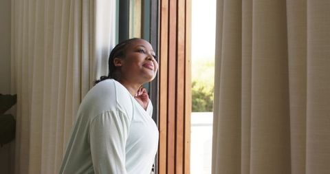 Mid-adult african american woman standing at sliding glass door enjoying sunlight and calm