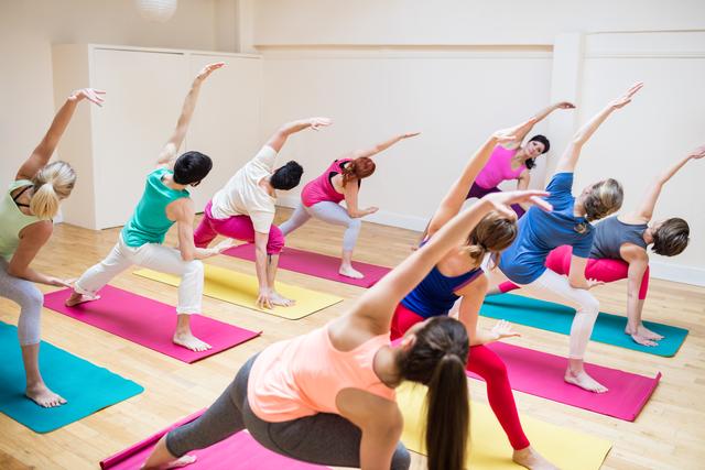 Group of people participating in a yoga class, guided by an instructor. They are performing stretching exercises on colorful mats in a bright, spacious studio. Ideal for promoting fitness classes, wellness programs, and healthy lifestyle activities.