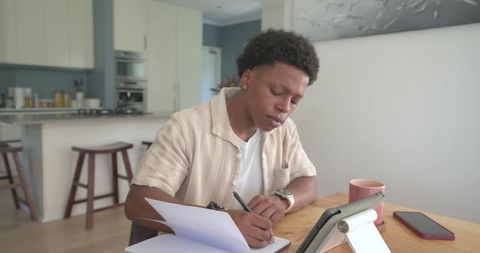 African American man writing in notebook at kitchen table with tablet stand, working remotely