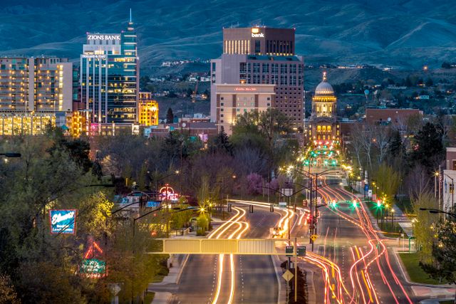 Image showcases an evening cityscape of Boise, Idaho with the Capitol Building prominently lit up. The busy streets capture the motion of city lights and activity. Useful for content related to Boise's tourism, business ads, urban life stories, and commercial district promotions.
