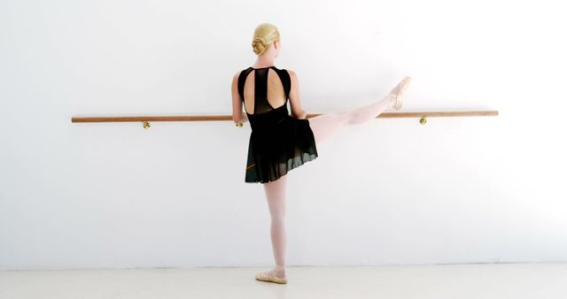 Elegant ballet dancer practicing leg stretches at a barre in dance studio, dressed in a black leotard and pointe shoes. Ideal for use in fitness, dance training, performing arts, and health and wellness promotion materials.