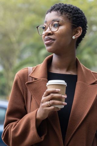 African woman standing holding takeaway coffee wearing brown coat glasses urban portrait