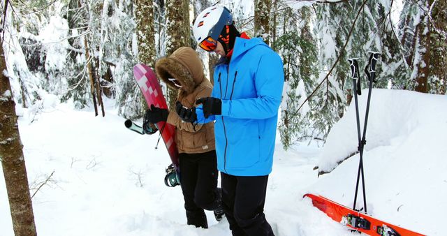 Two people, friends or a couple, are preparing for snowboarding in a snowy forest, with copy space. Wearing winter sports gear, the Caucasian man and woman are adjusting their equipment amidst the serene backdrop of snow-covered trees.