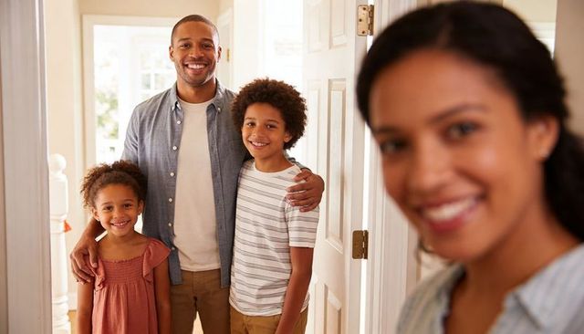Smiling family greeting at front door, parents welcoming kids into bright home foyer, in sunlight