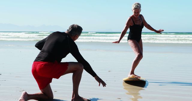 A senior Caucasian man is crouching on the sand as he supports a senior Caucasian woman attempting to balance on a surfboard at the beach, with copy space. Their playful activity suggests a moment of leisure and enjoyment in a coastal setting.