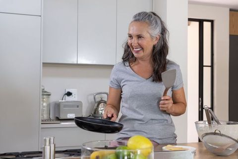 Mature woman cooking pancakes in modern kitchen smiling with spatula and pan