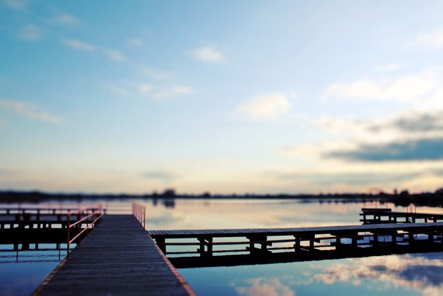 Peaceful view of a wooden pier extending into a calm lake at sunset, reflecting the soft evening light. Ideal for use in travel blogs, relaxation retreat promotions, nature-themed calendars, and landscape photography collections.