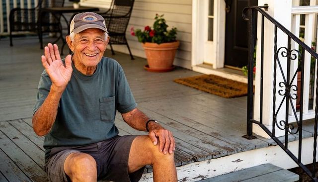 Smiling senior man waving and sitting on front porch steps wearing cap and wristwatch