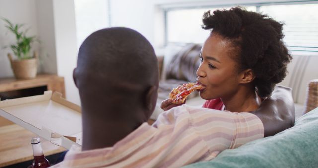 Image depicts a happy couple sitting on a couch, enjoying a slice of pizza in a modern living room. Perfect for use in lifestyle blogs, food delivery service advertisements, or promoting home dining and bonding moments.