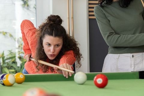 Female friends playing pool at home game room, woman aiming cue stick focusing on shot