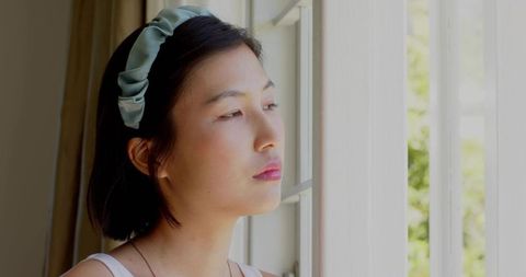 Korean woman gazing out window with pale green headband, contemplative daylight portrait